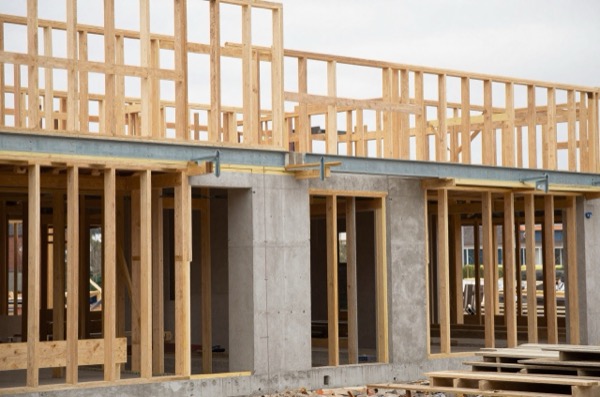 Residential building under construction with visible timber framing, steel beams, and concrete foundations.