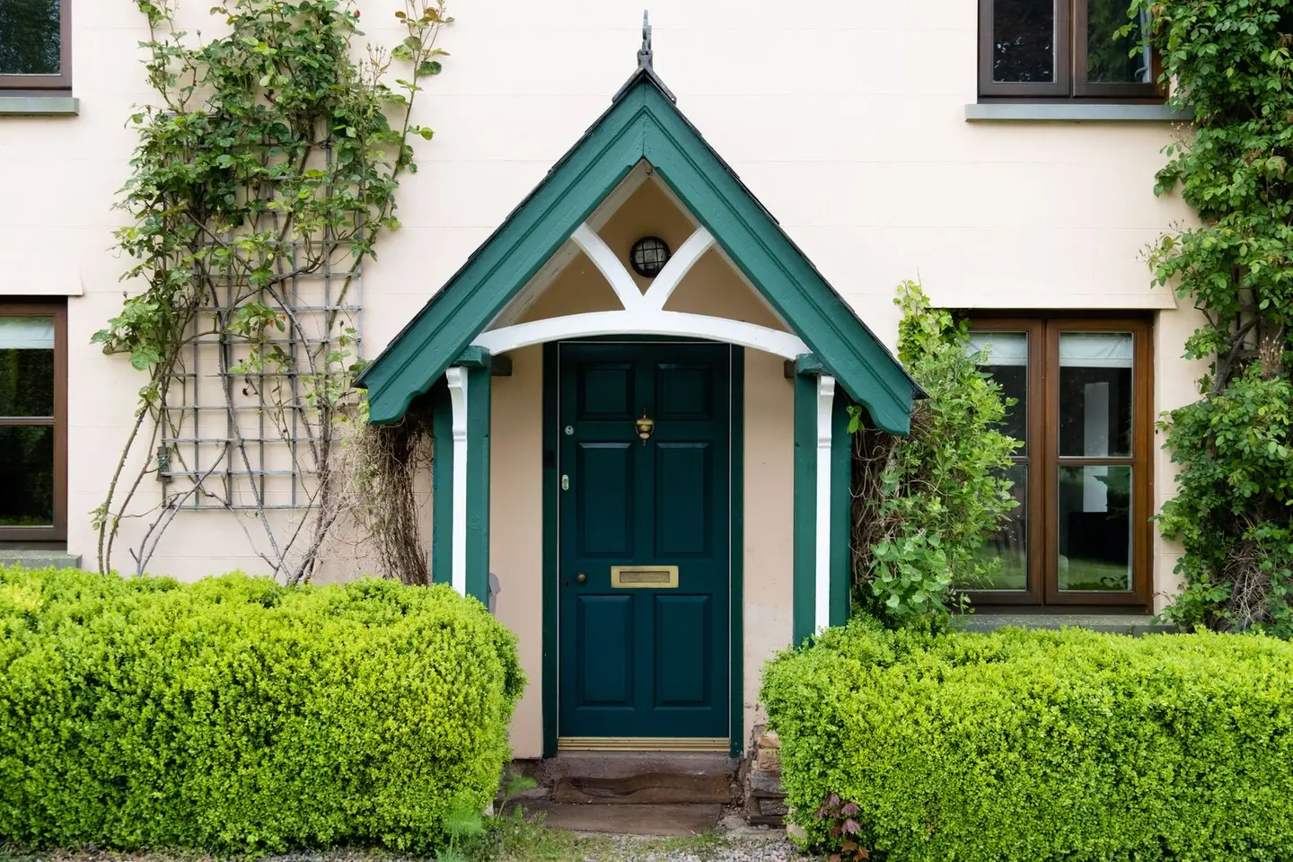 A pitched canopy porch over a teal front door on a rendered English cottage, with clipped hedges either side.