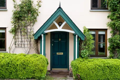 A pitched canopy porch over a teal front door on a rendered English cottage, with clipped hedges either side.