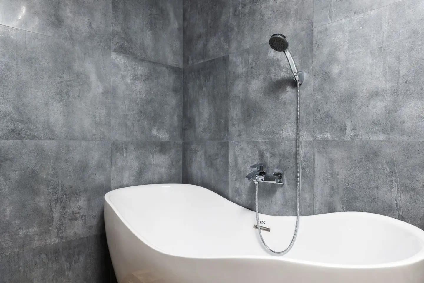 Interior of light modern bathroom with white bath under shower on grey tile wall