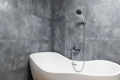 Interior of light modern bathroom with white bath under shower on grey tile wall