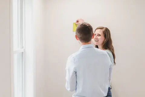 Man and woman choosing paint for home decor, holding colour swatch against wall.