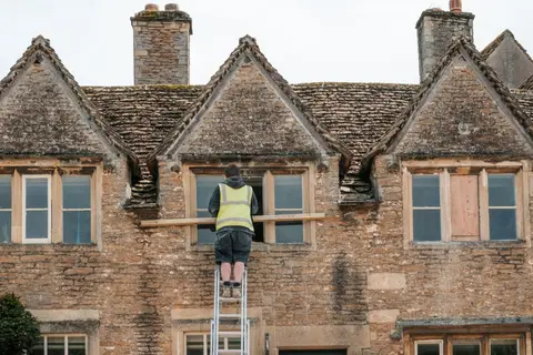 A construction worker on a ladder repairs a window on a stone building.