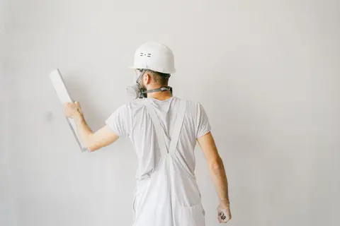 Back view of a construction worker in safety gear plastering a white wall.