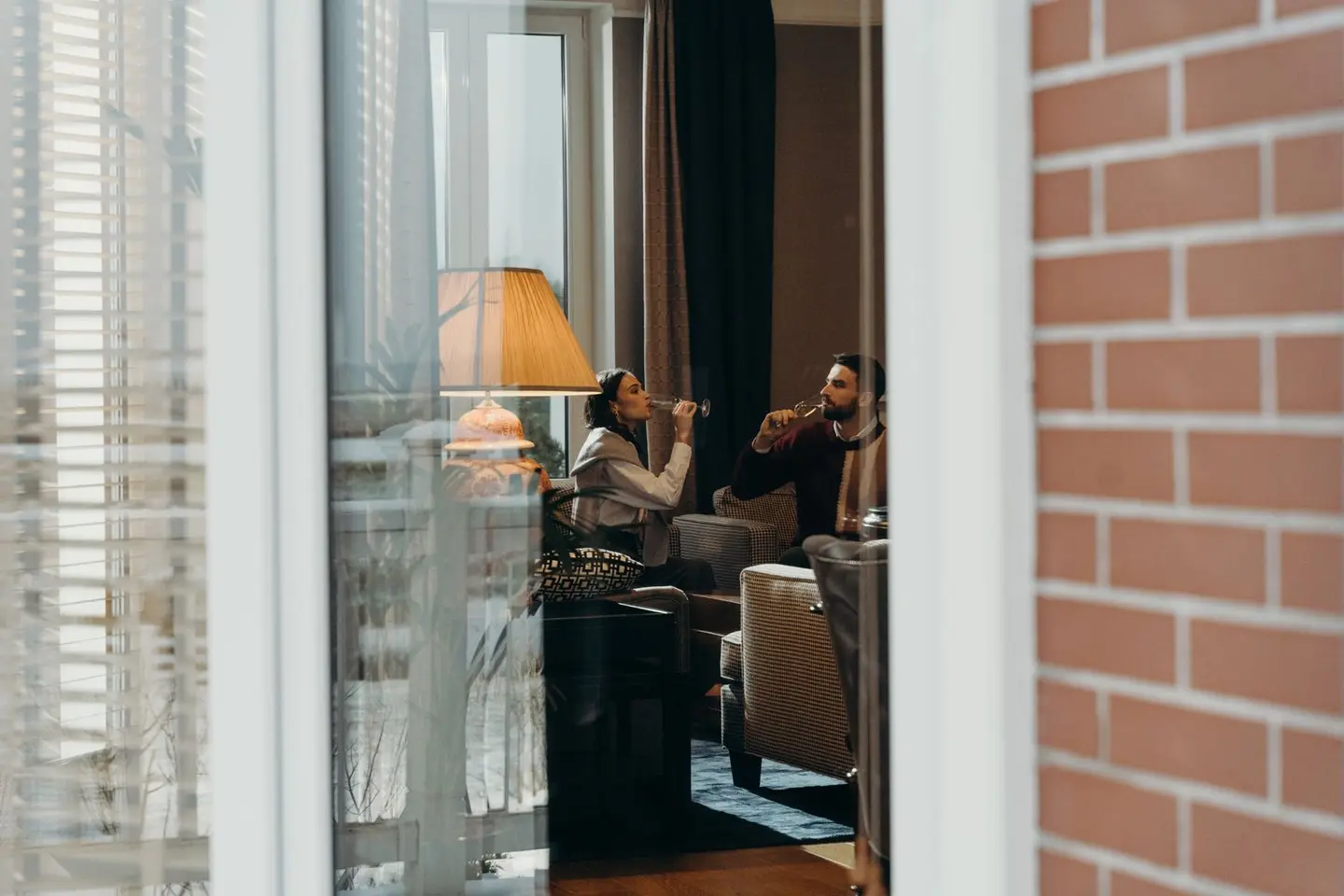 A couple relaxing and drinking in a modern, warm living room interior viewed through a window.