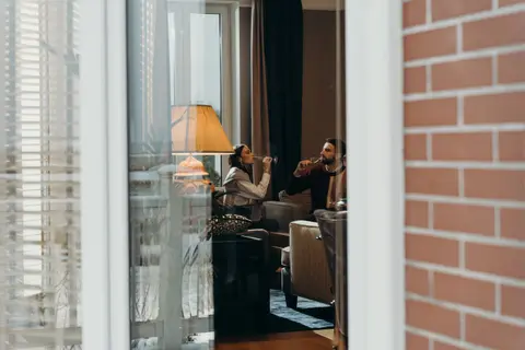 A couple relaxing and drinking in a modern, warm living room interior viewed through a window.
