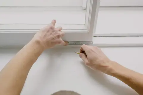 From above of crop unrecognizable male worker measuring window frame with metal ruler and pencil in house