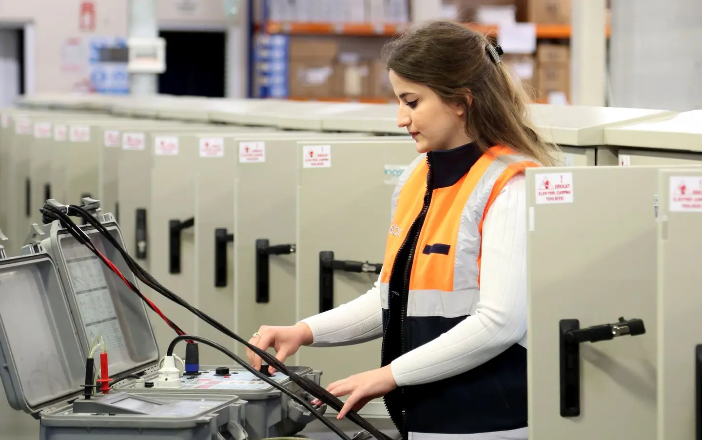 A female electrician in safety gear checks electrical panels indoors.