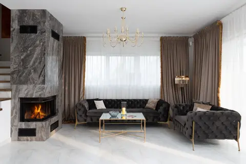 Interior of modern living room with soft couches and side table placed near windows with curtains and burning fireplace at home