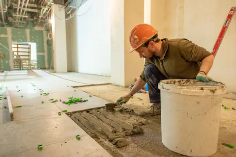 A construction worker lays tiles in an indoor renovation project, wearing safety gear.
