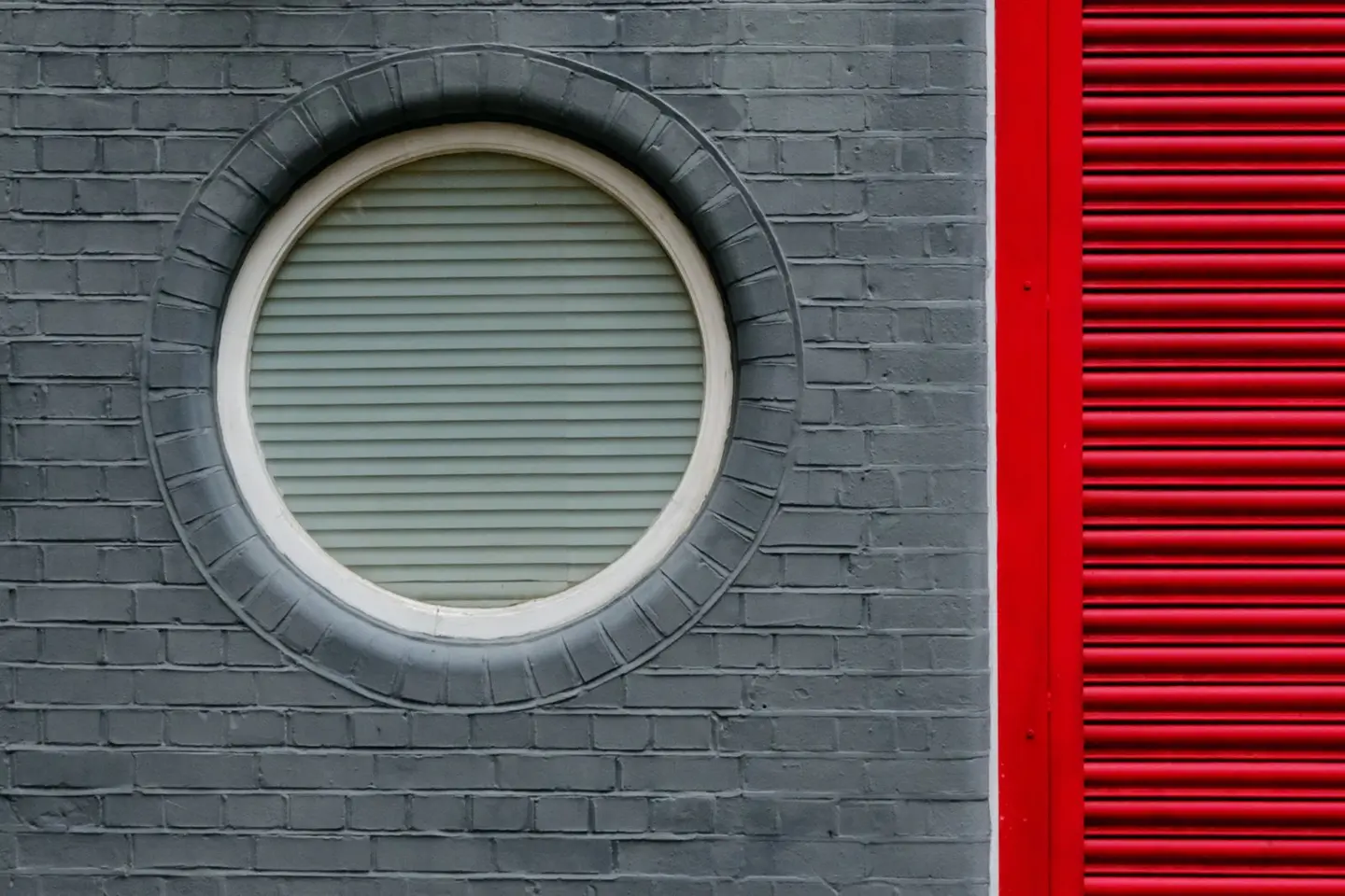 Unique architectural detail featuring a circular window on grey brick and red shutter. Captured in London.