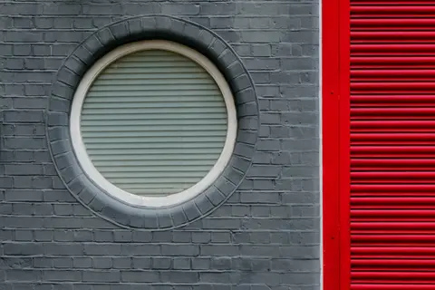 Unique architectural detail featuring a circular window on grey brick and red shutter. Captured in London.