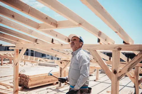 A construction worker surveys a wooden roof frame build on a sunny day.