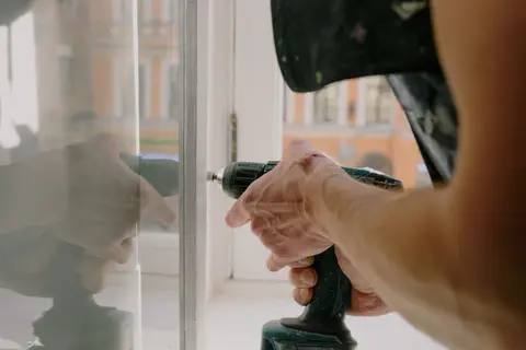 Unrecognizable professional male worker assembling white window with cordless screwdriver with drill near windowsill with reflection on glass during repair work