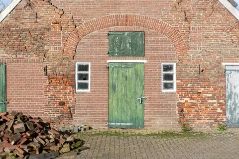 An aged brick farm building with green painted doors and cracked walls in sunlight.