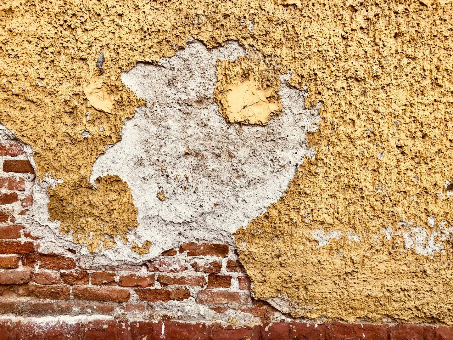 Close-up of a weathered wall showing exposed bricks and peeling plaster with a yellow hue.