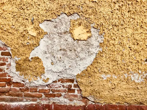 Close-up of a weathered wall showing exposed bricks and peeling plaster with a yellow hue.