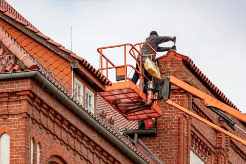 A roofer working from a cherry picker on the clay tiled roof of a red brick building.