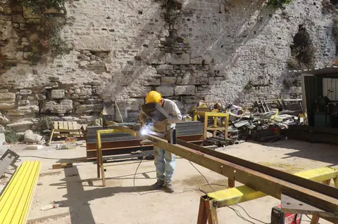 Worker in protective gear welding a steel beam at an outdoor construction site against a stone wall.