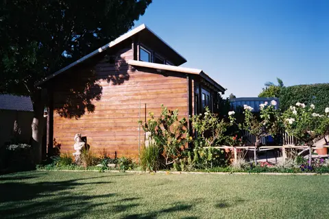 A rustic wooden house surrounded by a flourishing garden with roses and green grass under clear blue skies.