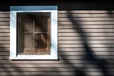 A rustic wooden house exterior showcasing a window with a mosquito net casting shadows in sunlight.